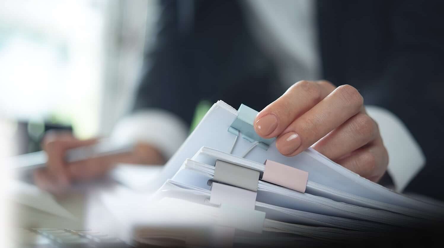 Eine Hand h&auml;lt einen Stapel von Dokumenten mit B&uuml;roklammern, w&auml;hrend eine andere Hand einen Stift h&auml;lt.