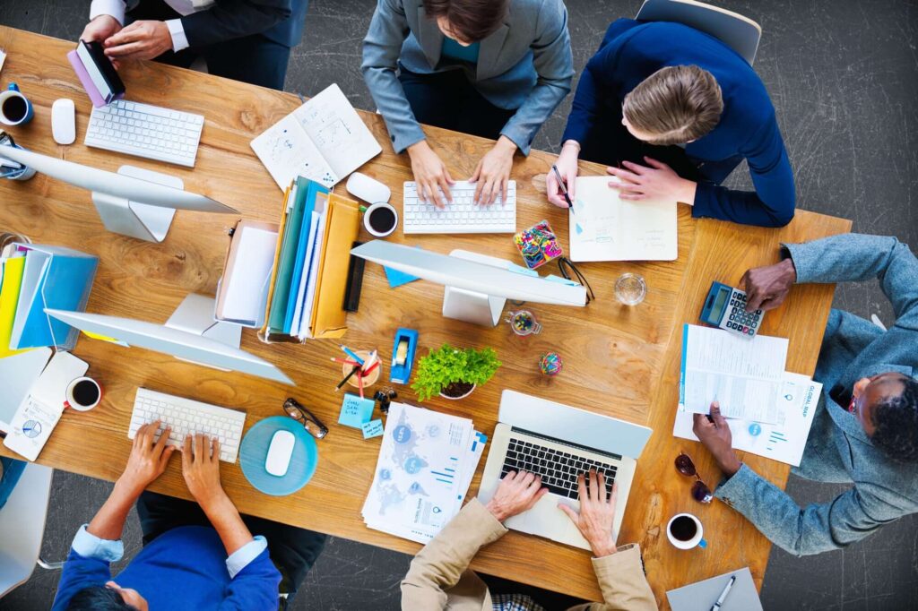 &Uuml;berblick &uuml;ber ein Teammeeting am Tisch mit Laptops, Notizen und Getr&auml;nken, umgeben von Unterlagen und B&uuml;romaterial.