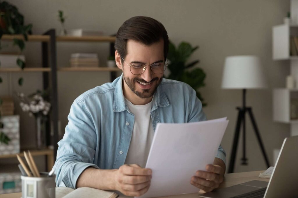 Mann mit Brille l&auml;chelt, w&auml;hrend er Dokumente am Schreibtisch in einem modernen Raum mit Pflanzen und Laptop betrachtet.