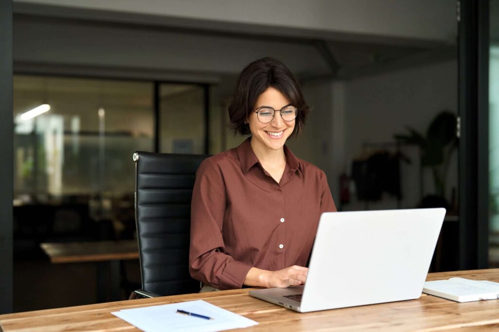Eine Frau mit Brille sitzt an einem Schreibtisch und arbeitet l&auml;chelnd an einem Laptop.