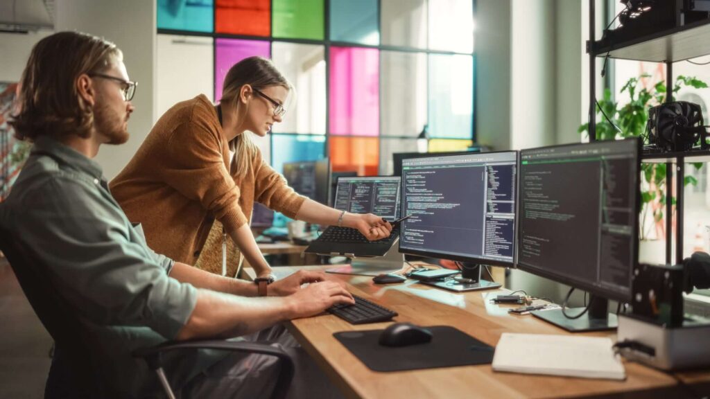 Zwei Personen in einem modernen B&uuml;ro arbeiten an Computern mit Code auf mehreren Bildschirmen.