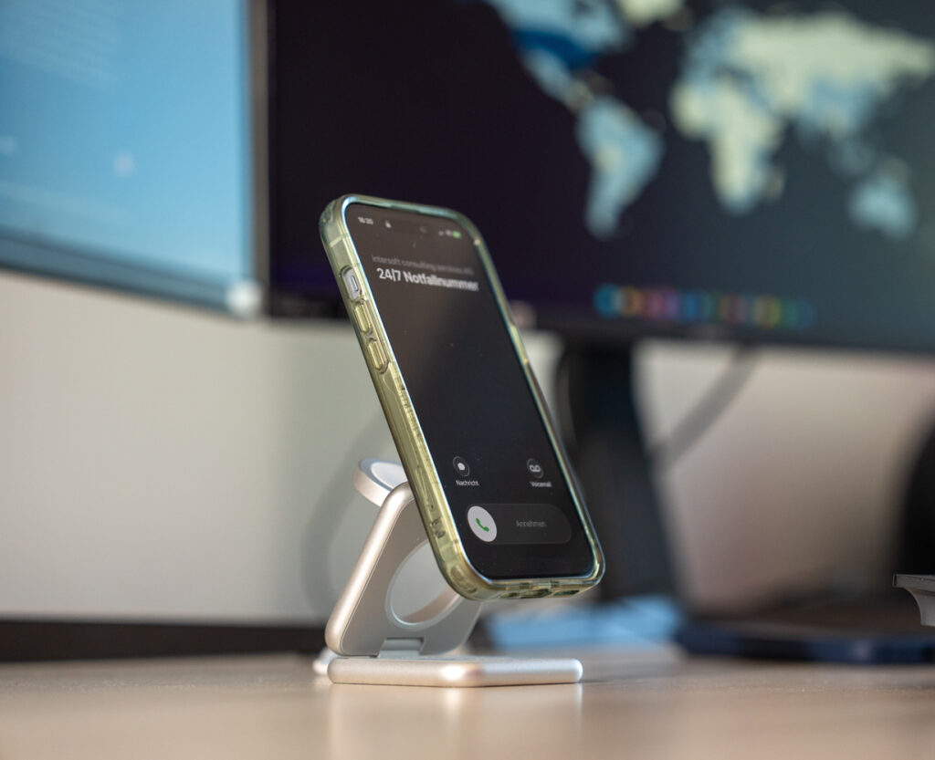Ein Smartphone steht auf einem silbernen St&auml;nder, sichtbar ist ein Anrufbildschirm mit der Aufschrift &bdquo;24/7 Verf&uuml;gbarkeit&ldquo;.