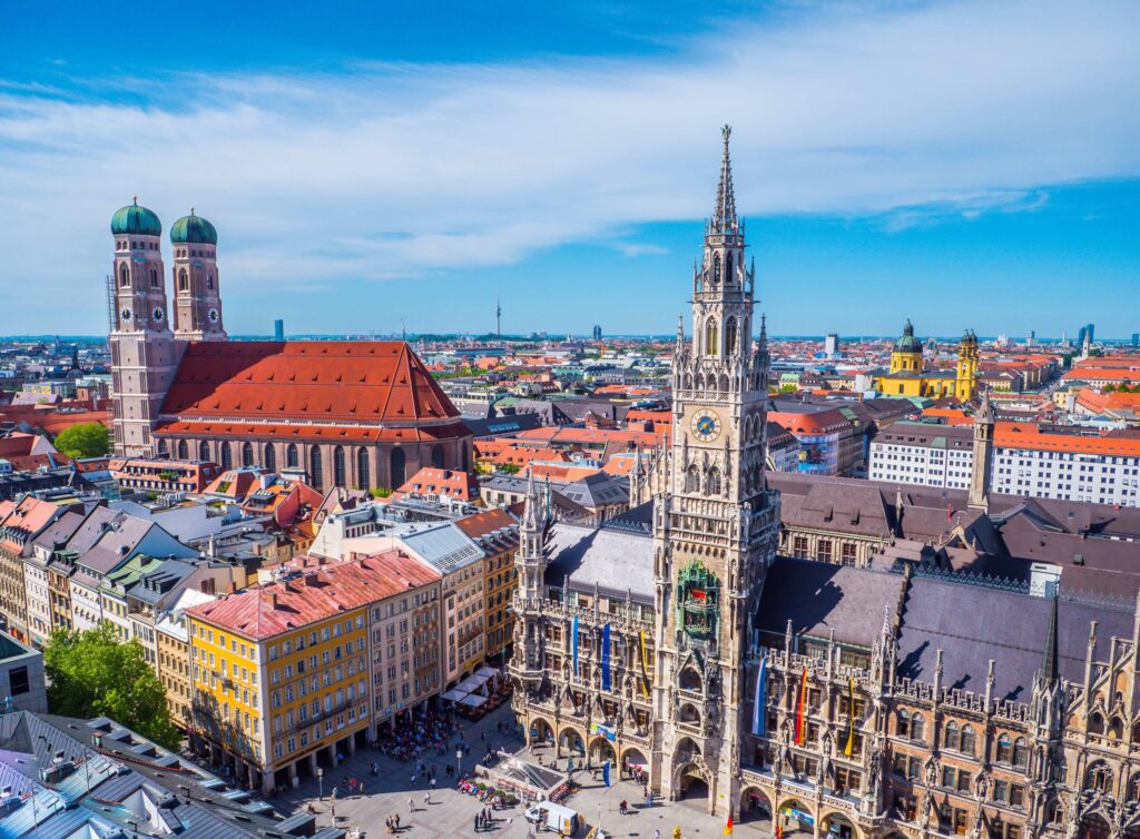 Blick auf M&uuml;nchens Marienplatz mit dem Neuen Rathaus und der Frauenkirche im Hintergrund unter blauem Himmel.