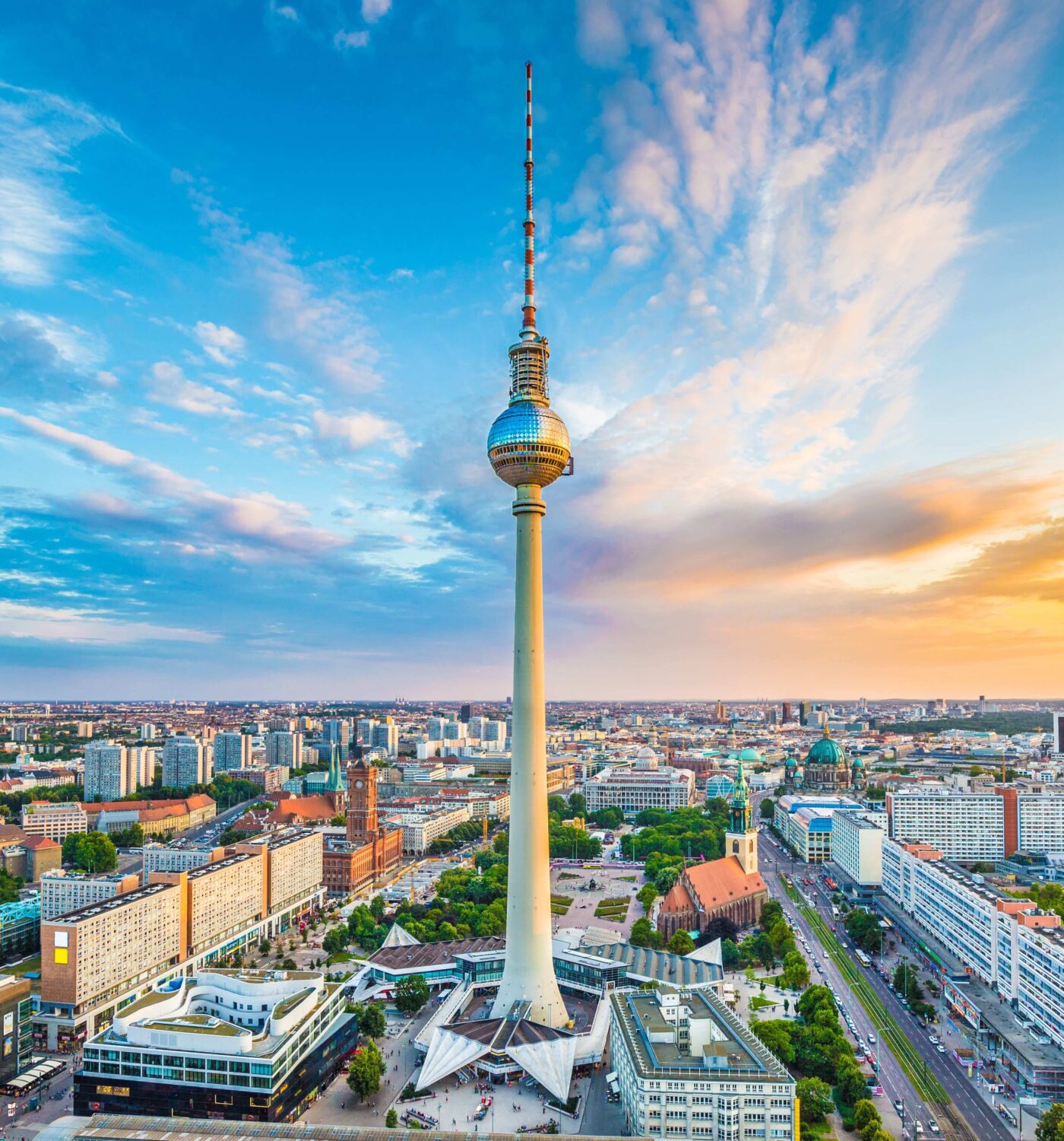 Fernsehturm Berlin bei Sonnenuntergang, umgeben von Stadtlandschaft und Wolken am Himmel.