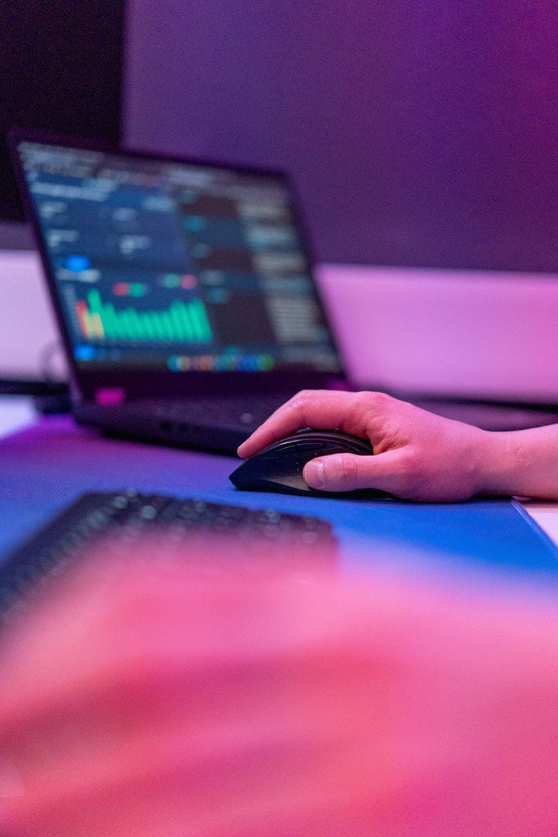 Eine Hand h&auml;lt eine Maus, w&auml;hrend ein Laptop mit bunten Grafiken im Hintergrund zu sehen ist.