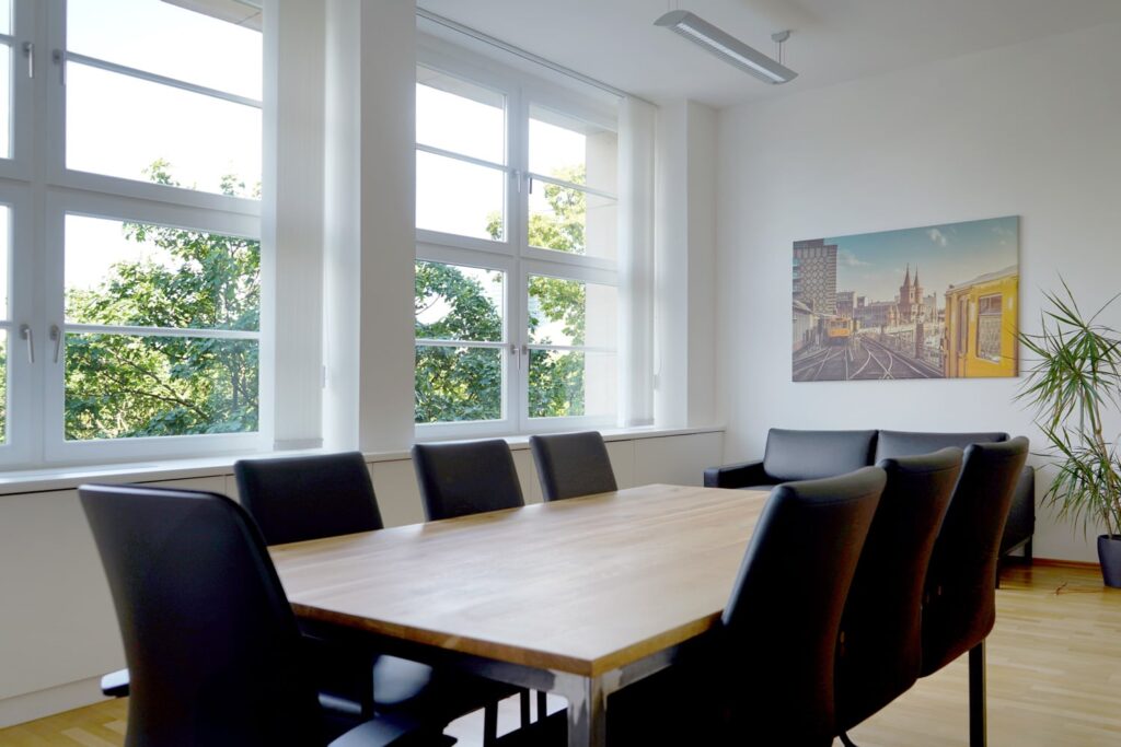 Ein moderner Besprechungsraum mit einem langen Holztisch, schwarzen St&uuml;hlen und gro&szlig;en Fenstern, die viel Tageslicht bieten.