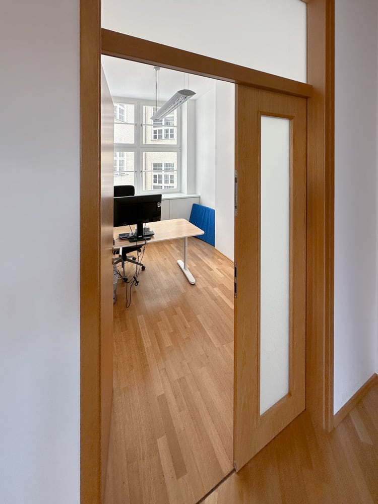 B&uuml;ro mit Holzt&uuml;r, Schreibtisch, Stuhl und Computer, helles Licht durch Fenster, moderne Einrichtung.