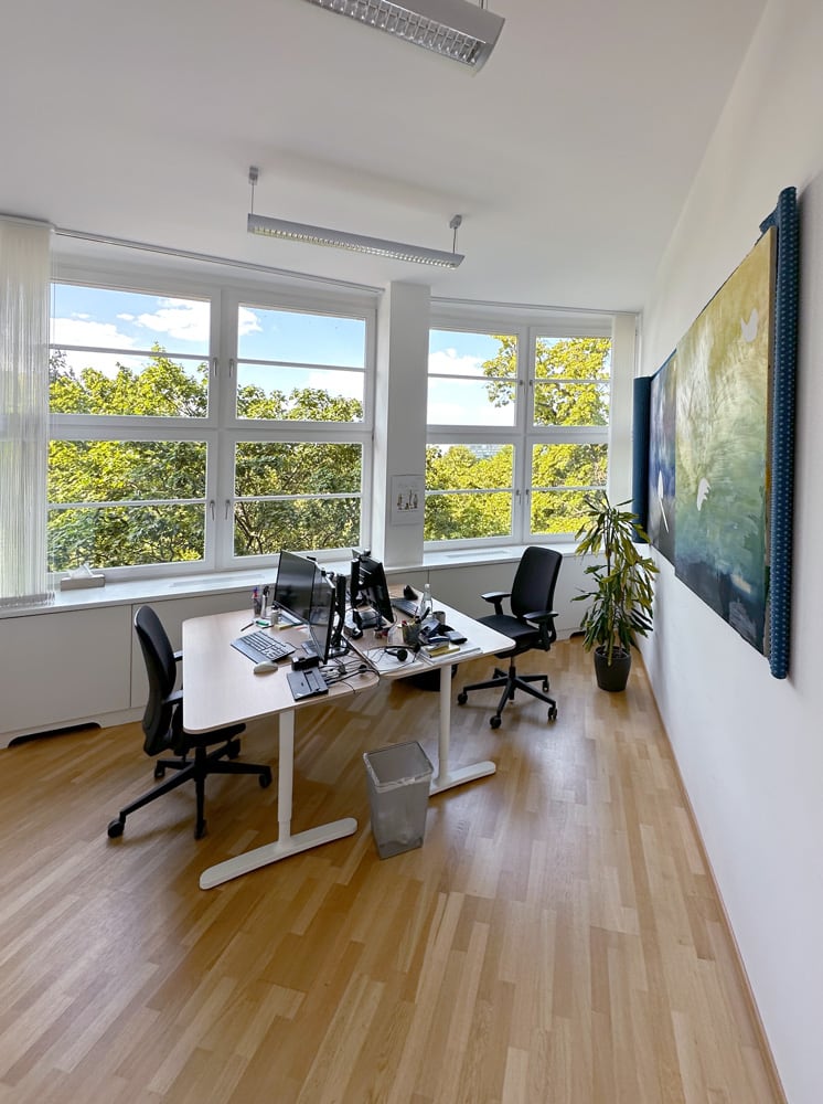 B&uuml;ro mit zwei Schreibtischen, Computerarbeitspl&auml;tzen und gro&szlig;en Fenstern mit Blick ins Gr&uuml;ne.