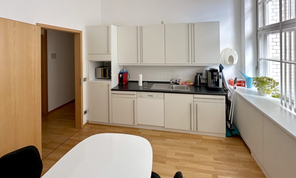 Moderne K&uuml;che mit wei&szlig;en Schr&auml;nken, Sp&uuml;le, Mikrowelle und Kaffeemaschine, Holzfu&szlig;boden und gro&szlig;en Fenstern.