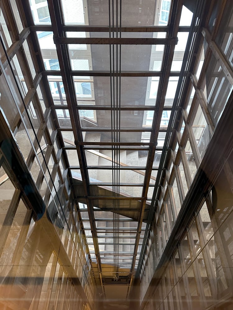 Blick nach unten in einen modernen Aufzugschacht mit Glasw&auml;nden und metallenen F&uuml;hrungsschienen.