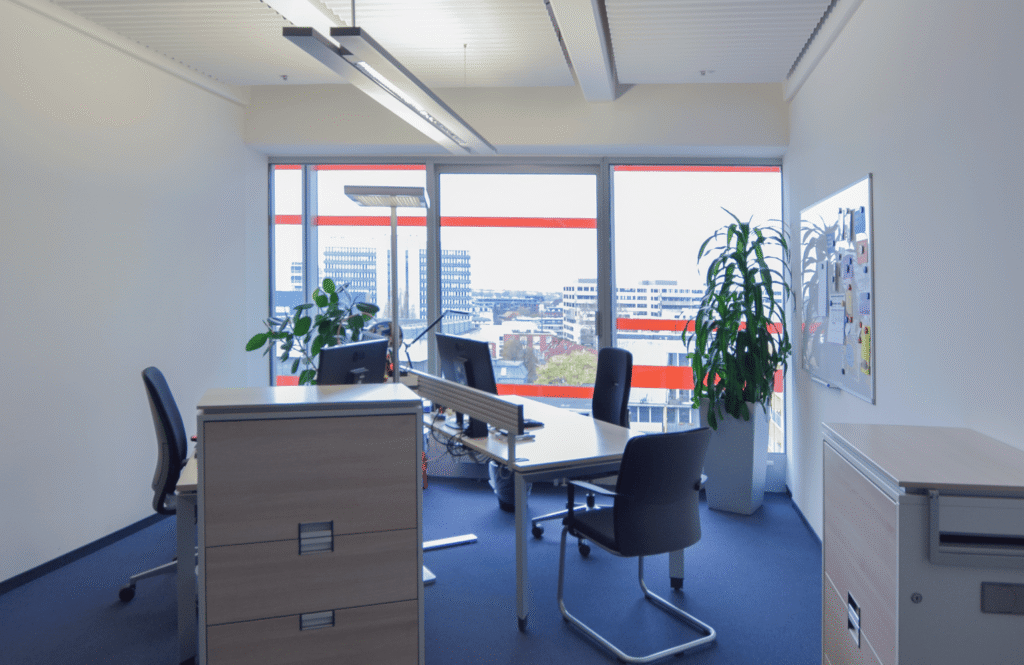 Ein modernes B&uuml;ro mit zwei Schreibtischen, St&uuml;hlen, Pflanzen und gro&szlig;en Fenstern mit Blick auf eine Stadtlandschaft.