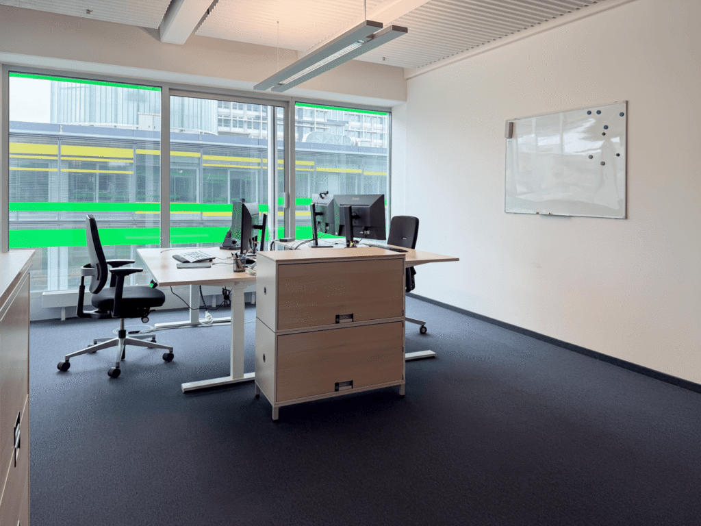 B&uuml;ro mit zwei Schreibtischen, St&uuml;hlen, Computern und einem wei&szlig;en Board, helle Fenster mit gr&uuml;nen Akzenten.
