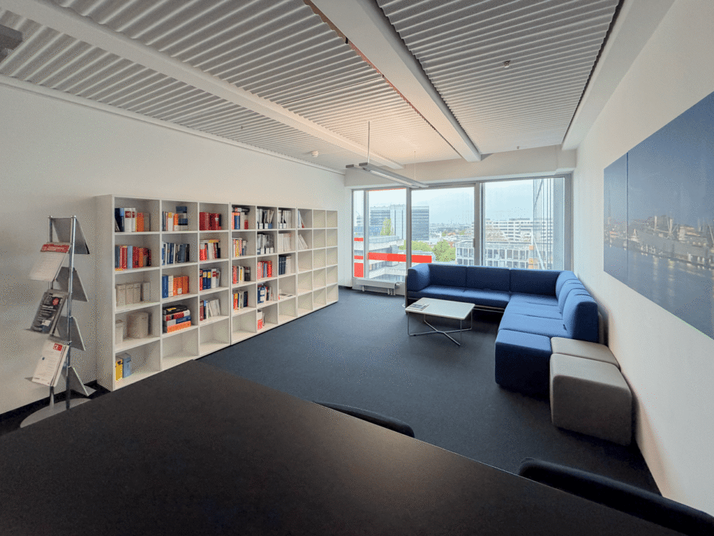 Heller Besprechungsraum mit B&uuml;cherregal, blauer Couch und Tisch, Blick auf die Stadt durch gro&szlig;e Fenster.