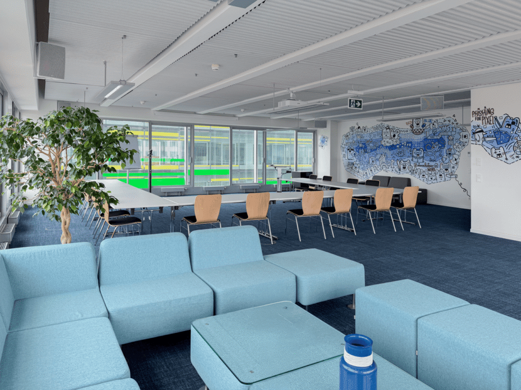 Moderner B&uuml;roraum mit blauen Sofas, einem langen Tisch, einem Pflanzenarrangement und einer bunten Wandgrafik.