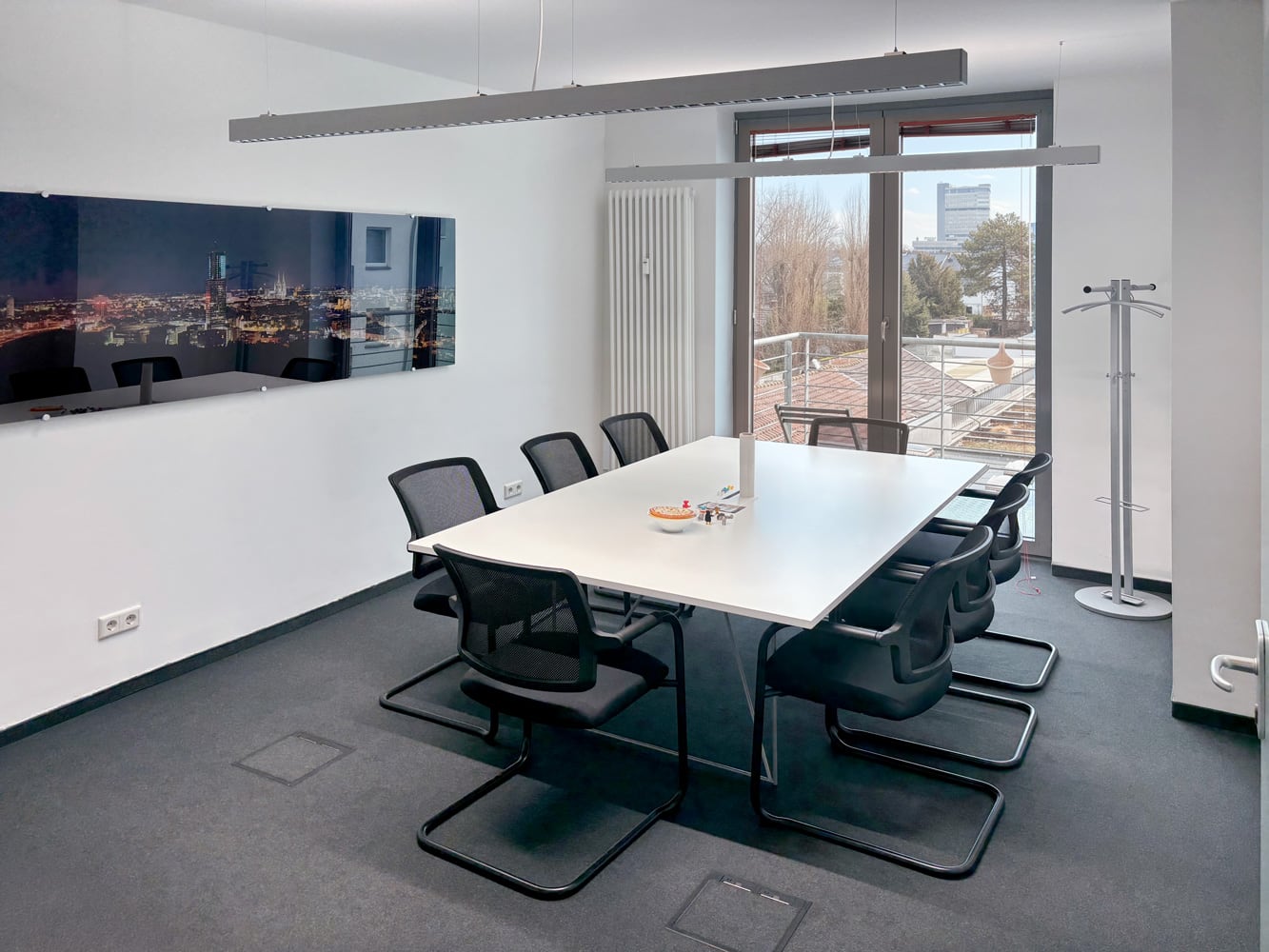 Ein moderner Besprechungsraum mit einem gro&szlig;en Tisch, acht St&uuml;hlen und Fensterblick auf eine Stadtlandschaft.
