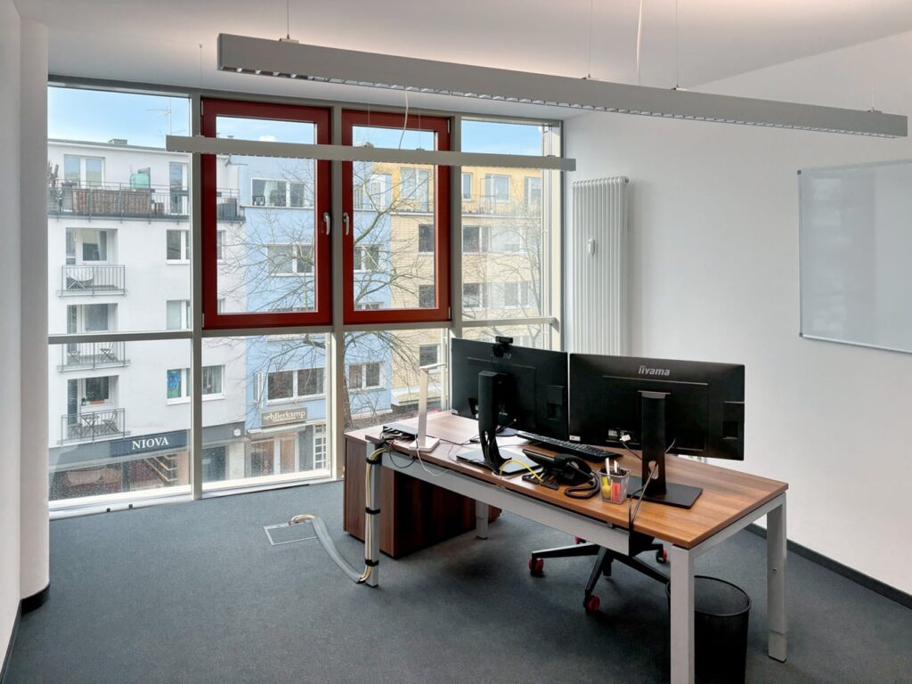 Ein moderner B&uuml;roraum mit Schreibtisch, zwei Monitoren und gro&szlig;en Fenstern, die auf ein st&auml;dtisches Umfeld blicken.