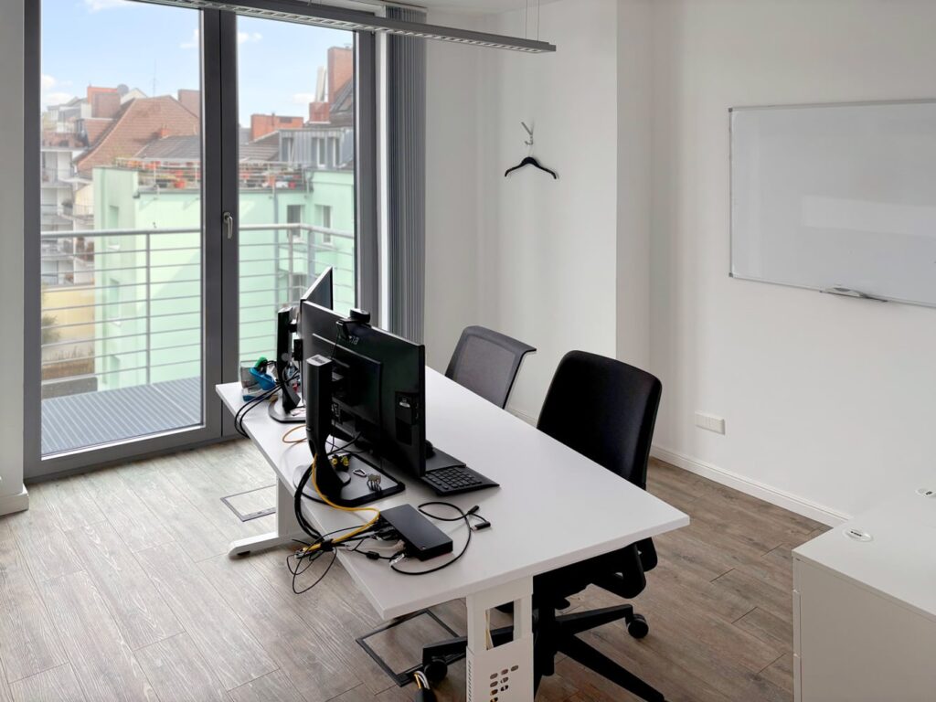 Ein moderner B&uuml;roraum mit zwei Computerbildschirmen, einem Schreibtisch, einem Stuhl und gro&szlig;en Fenstern.
