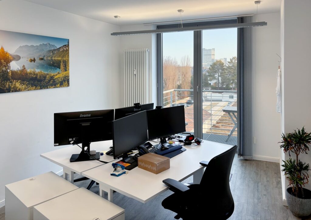 Modernes B&uuml;ro mit zwei Bildschirmen, Schreibtisch, Pflanzen und Blick auf einen Balkon mit Aussicht auf die Stadt.