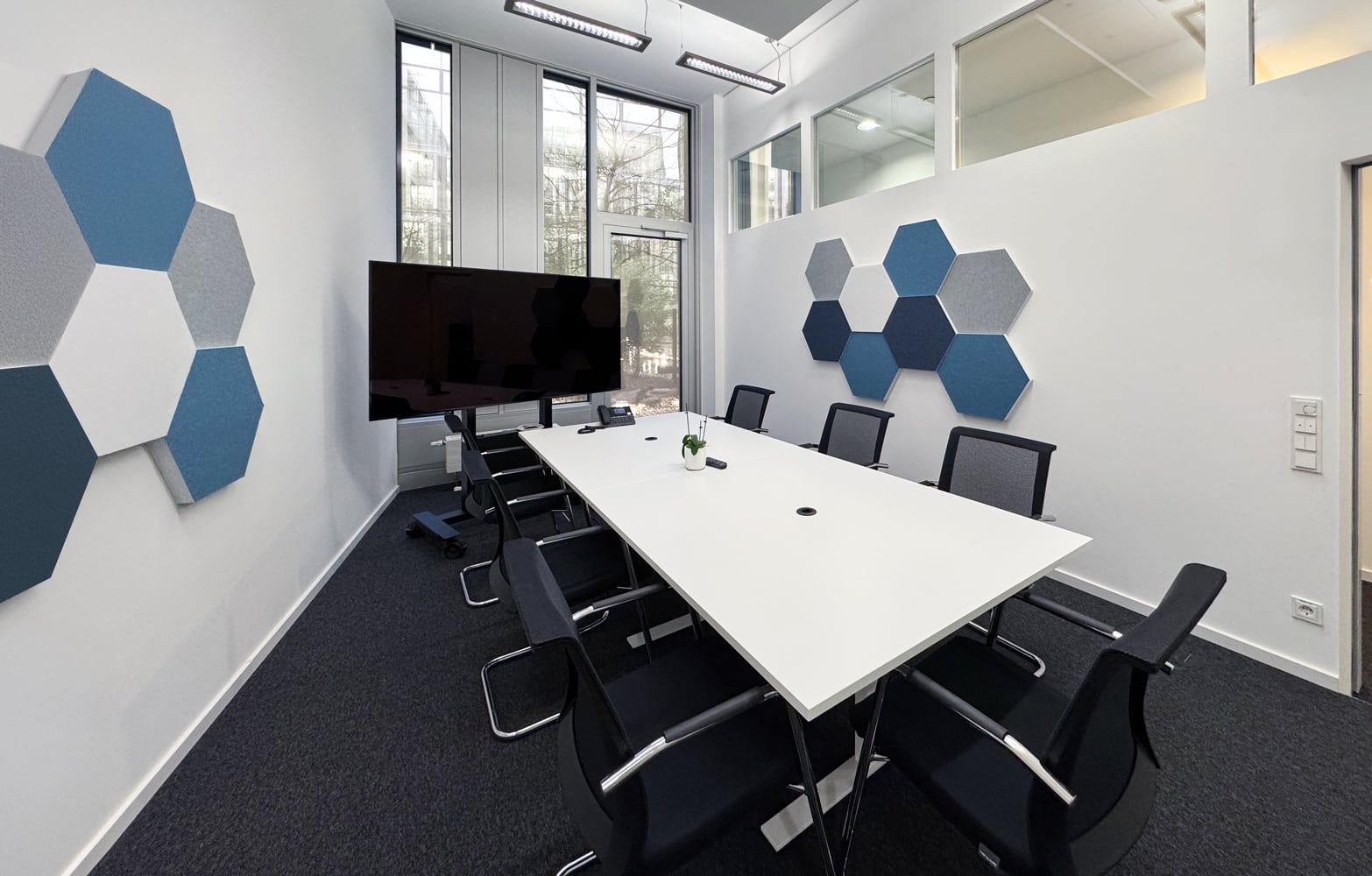 Moderne Besprechungsraum mit hexagonalen Wandpaneelen, einem gro&szlig;en Tisch und St&uuml;hlen, ausgestattet mit einem Bildschirm.