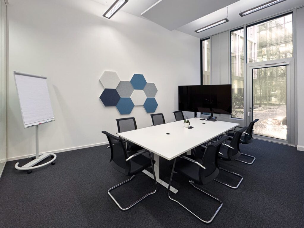 Ein moderner Konferenzraum mit einem gro&szlig;en Tisch, St&uuml;hlen, einem Bildschirm und hexagonalen Wandpaneelen.