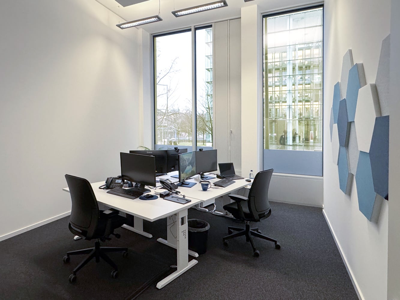 B&uuml;ro mit zwei Schreibtischen, je zwei Monitoren, B&uuml;rost&uuml;hlen und gro&szlig;en Fenstern mit Blick auf eine moderne Architektur.