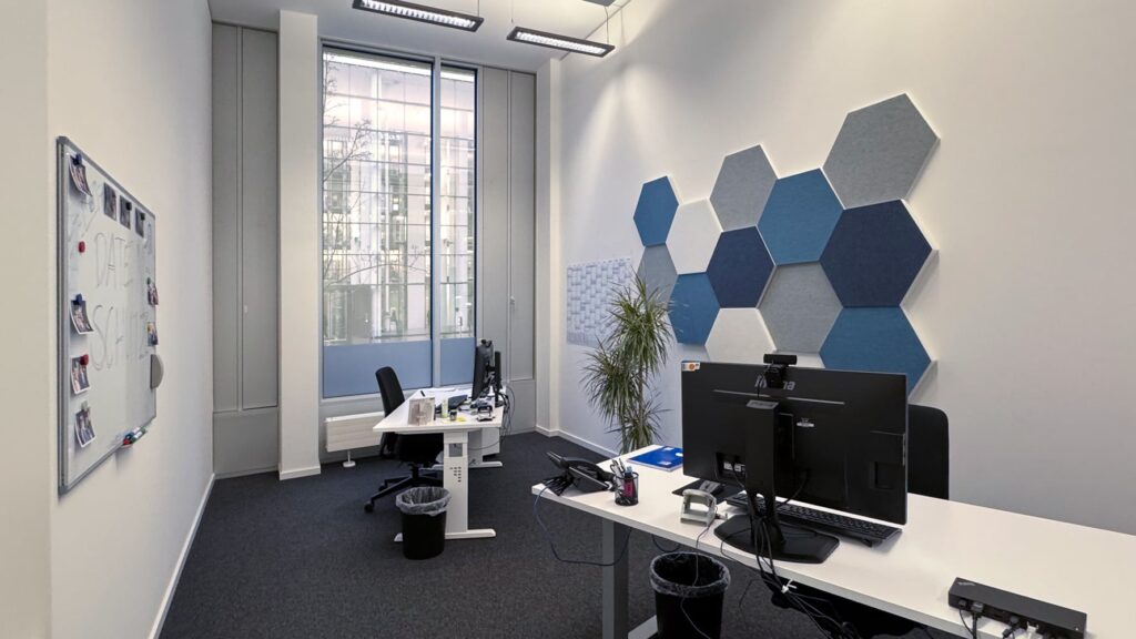 Ein modernes B&uuml;ro mit zwei Schreibtischen, einem gro&szlig;en Fenster und einer hexagonalen Wanddekoration in Blau- und Graut&ouml;nen.