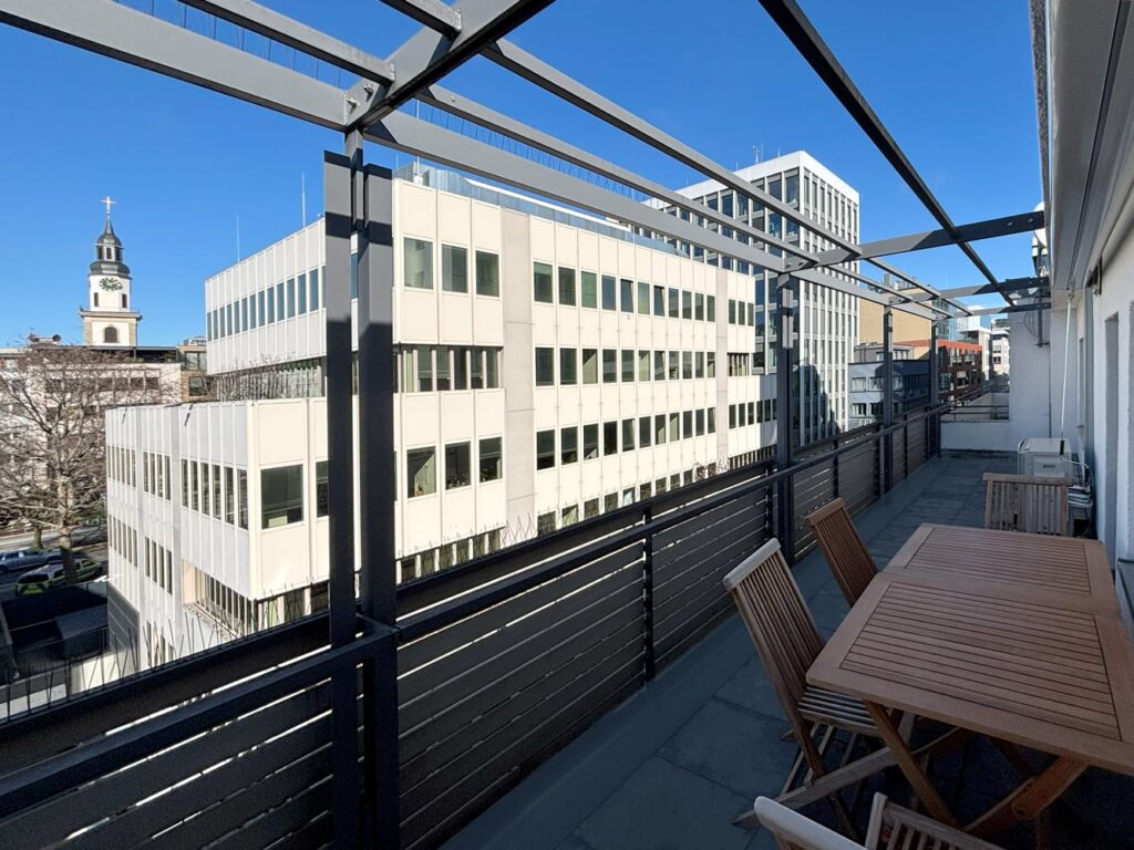 Blick von einem Balkon auf moderne Geb&auml;ude und einen Kirchturm bei klarem Himmel.