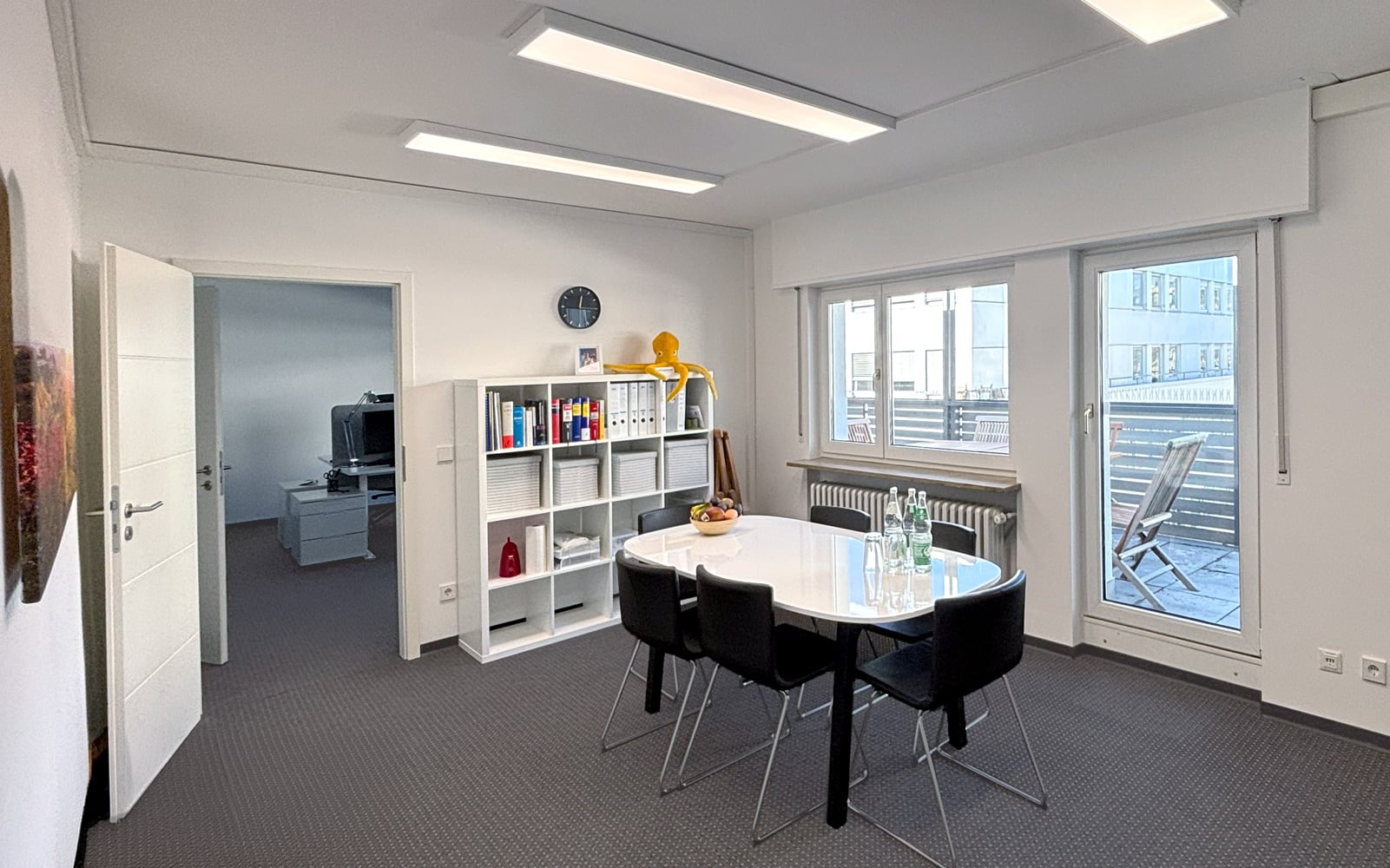 Modernes B&uuml;ro mit runder Tischgruppe, B&uuml;cherregal und Zugang zu einem Balkon. Helle W&auml;nde und gro&szlig;e Fenster.
