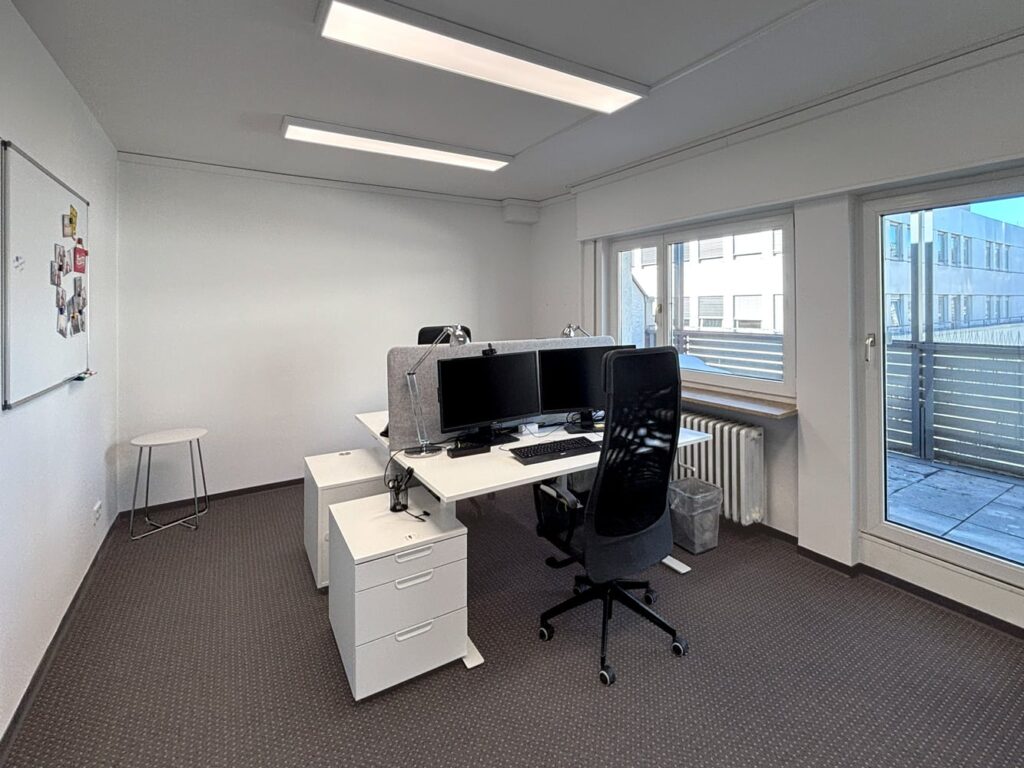 B&uuml;ro mit zwei Schreibtischen, zwei Monitoren und einem Fenster, das auf einen Balkon mit Sicht nach au&szlig;en zeigt.