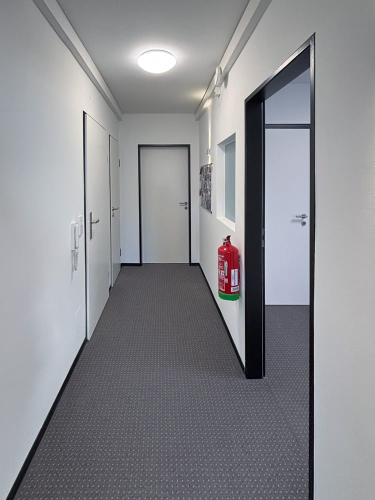 Ein schmaler Flur mit wei&szlig;en W&auml;nden, grauem Teppichboden und einer roten Feuerl&ouml;scher an der Wand.
