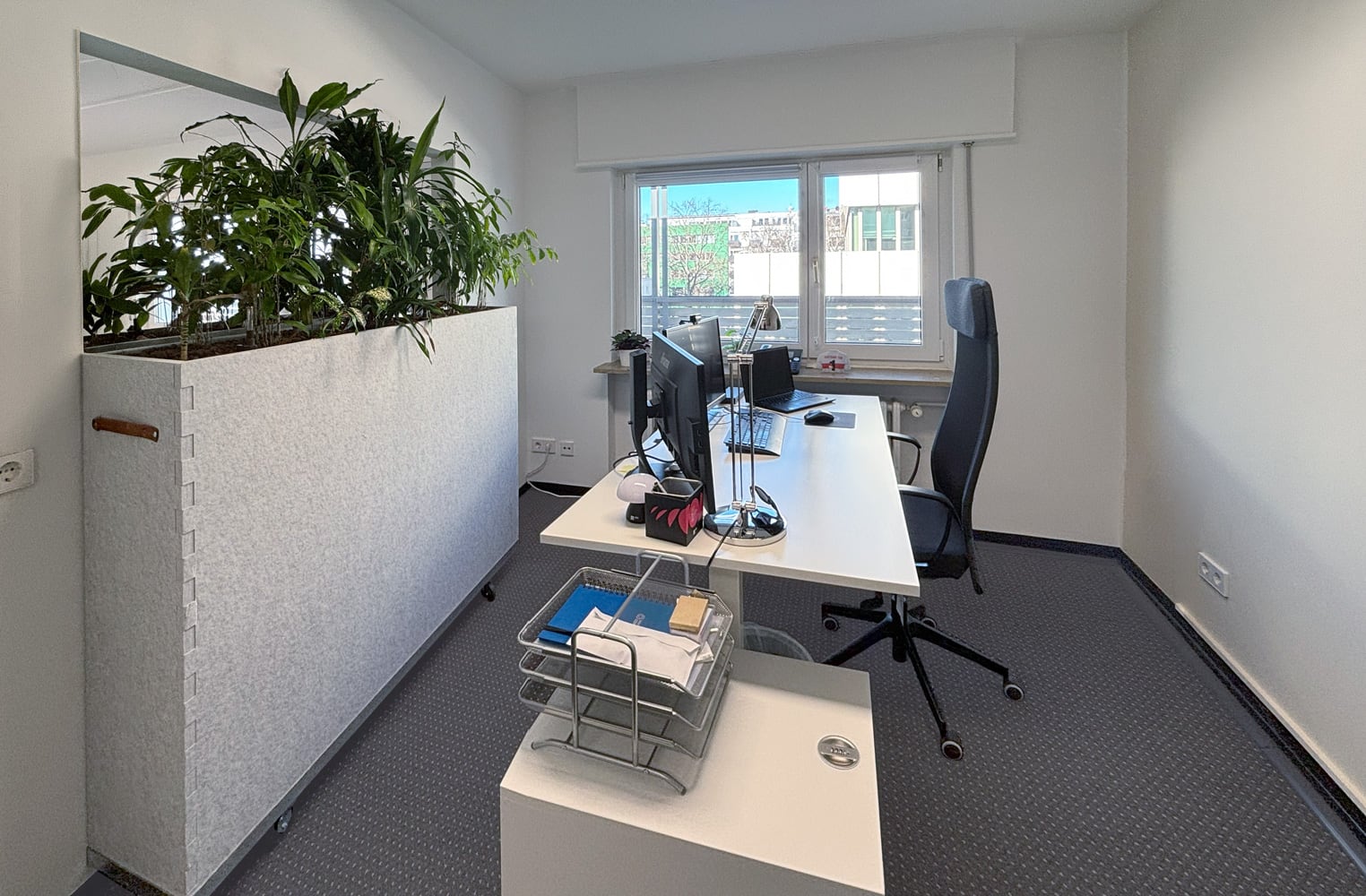 Ein modernes B&uuml;ro mit Schreibtisch, Stuhl, Pflanzenwand und gro&szlig;en Fenstern, die viel Tageslicht hereinlassen.