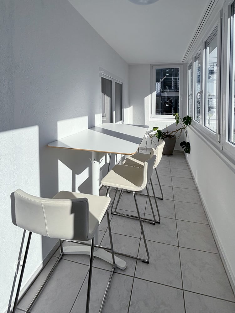Heller, moderner Raum mit einem langen Tisch und vier wei&szlig;en St&uuml;hlen, umgeben von Fenstern und Pflanzen.