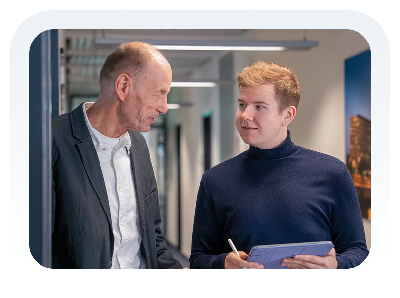 Zwei Männer im Gespräch im Büroflur, einer hält ein Tablet, der andere hört aufmerksam zu.